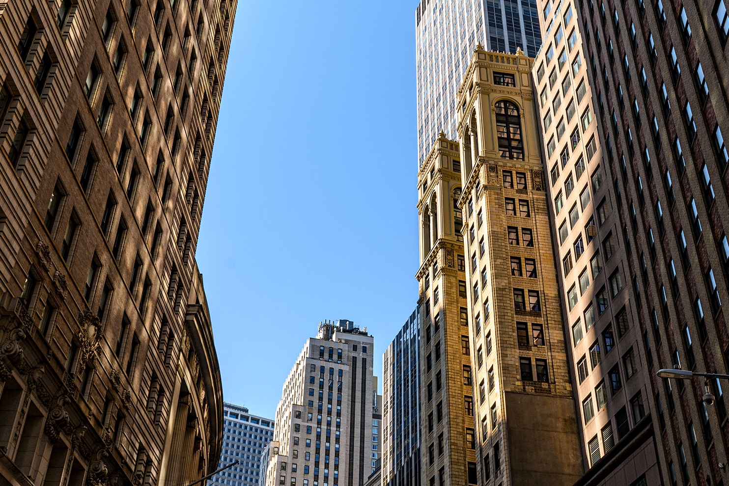 A view from the street looking up at tall, historic skyscrapers in a city. The buildings feature intricate architectural details, with a bright blue sky in the background.