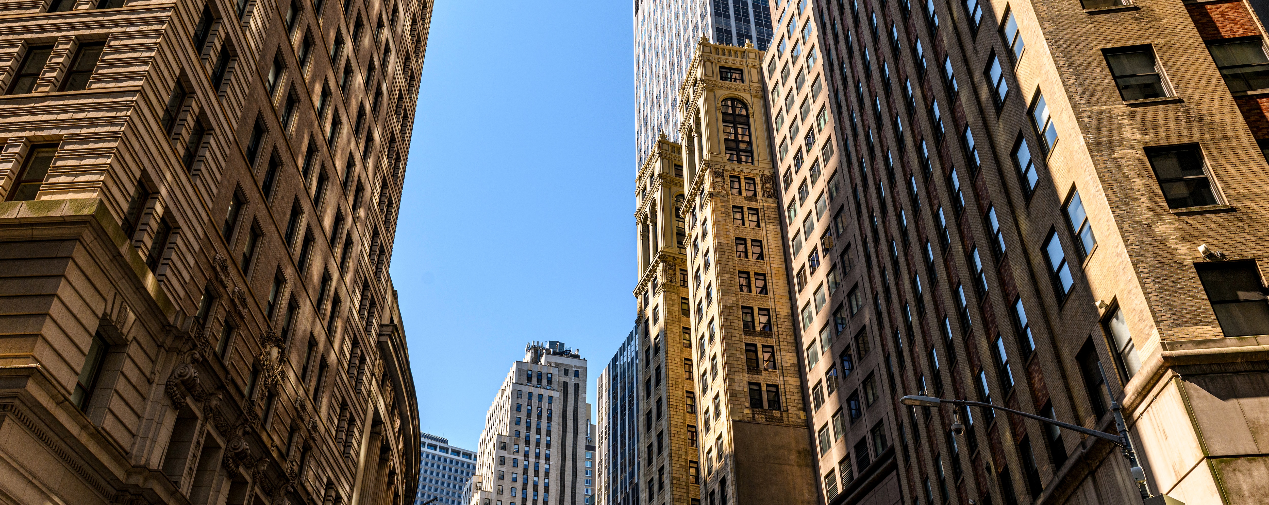 A view from the street looking up at tall, historic skyscrapers in a city. The buildings feature intricate architectural details, with a bright blue sky in the background.