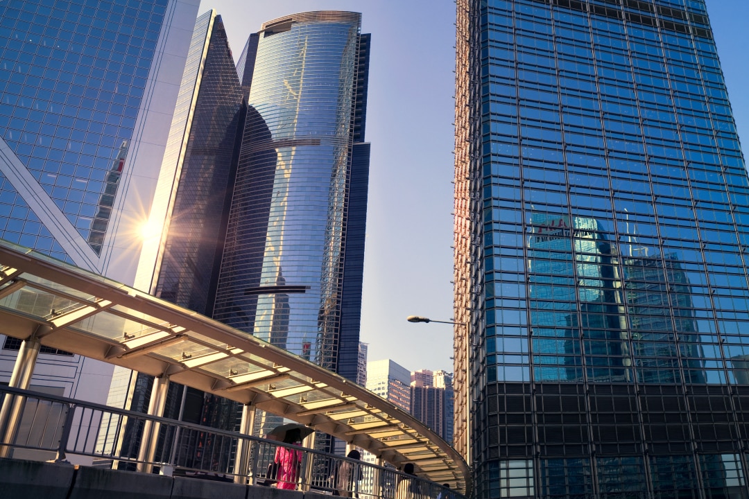 A view from the street looking up at tall, historic skyscrapers in a city. The buildings feature intricate architectural details, with a bright blue sky in the background.