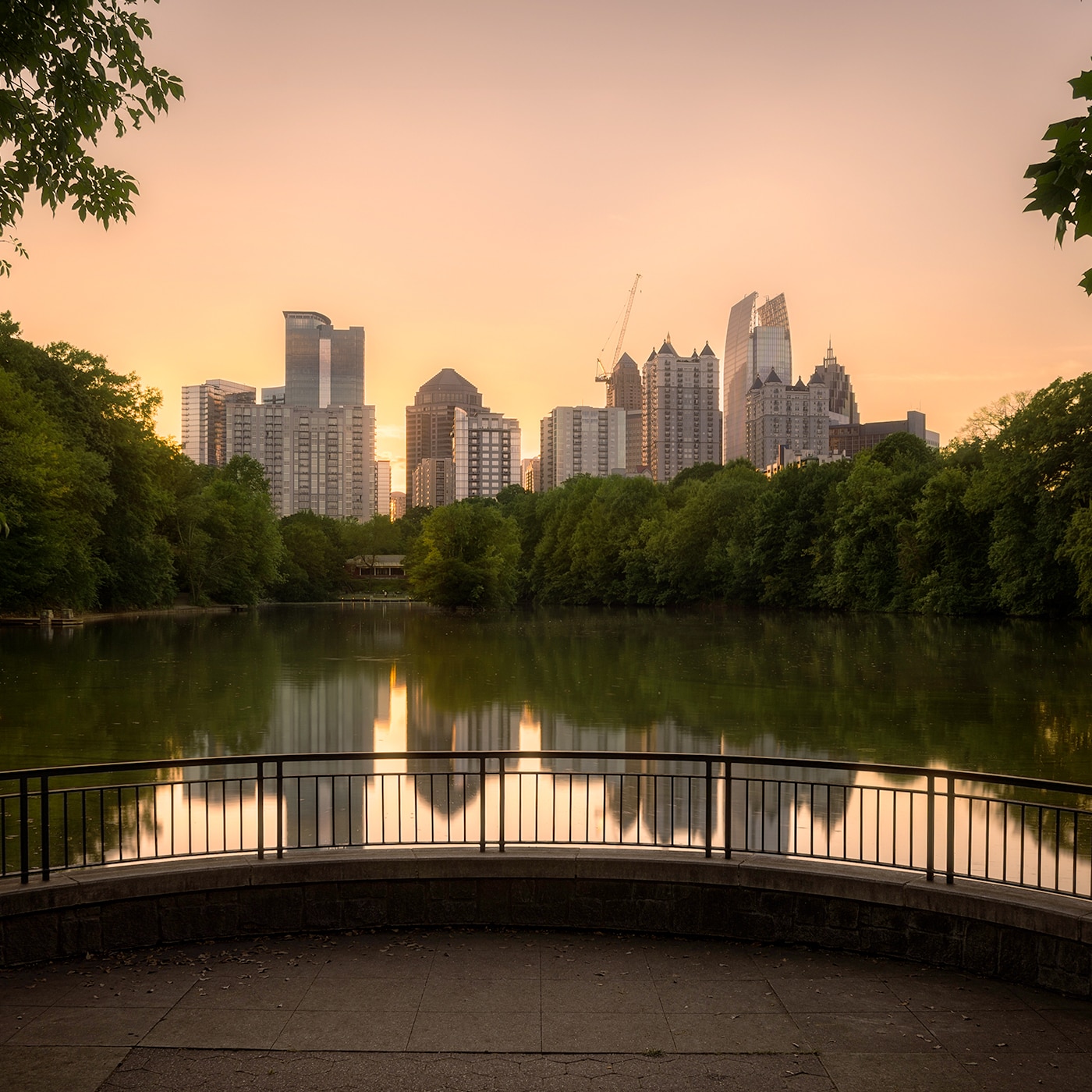 A serene cityscape view at sunset, featuring a skyline of modern high-rise buildings reflecting in a calm lake. Lush green trees frame the image, and a curved railing in the foreground adds depth. The warm hues of the sky contrast with the cool tones of the water and greenery