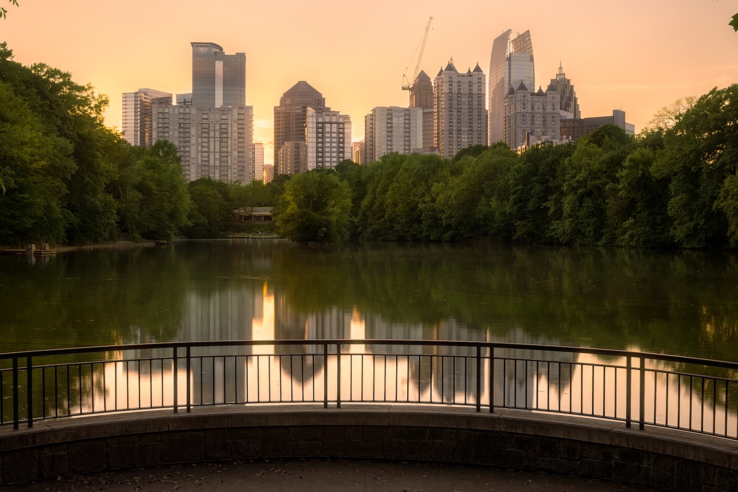 A serene cityscape view at sunset, featuring a skyline of modern high-rise buildings reflecting in a calm lake. Lush green trees frame the image, and a curved railing in the foreground adds depth. The warm hues of the sky contrast with the cool tones of the water and greenery