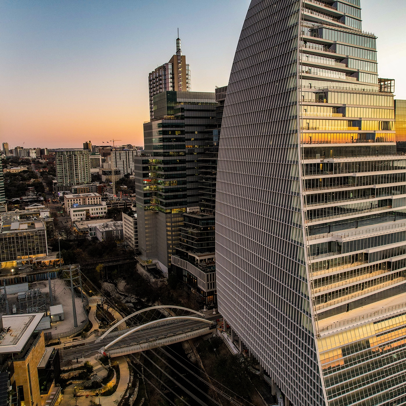 A modern cityscape at sunset featuring a striking curved glass skyscraper reflecting the warm hues of the sky. Surrounding high-rise buildings add to the urban density, while a sleek pedestrian bridge spans a railway below. The city extends into the distance with a mix of contemporary and older architecture