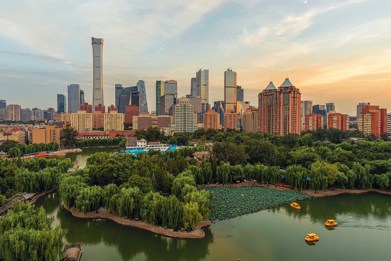 A scenic view of a lush urban park with a green lake, small islands, and traditional Chinese architecture, set against the modern skyline of Beijing at sunset. Yellow paddle boats float on the water, surrounded by dense trees and willows. The city's skyscrapers, including the distinctive China Zun Tower, rise in the background, showcasing a blend of nature and contemporary urban development. The soft hues of the sky enhance the tranquil yet vibrant atmosphere.