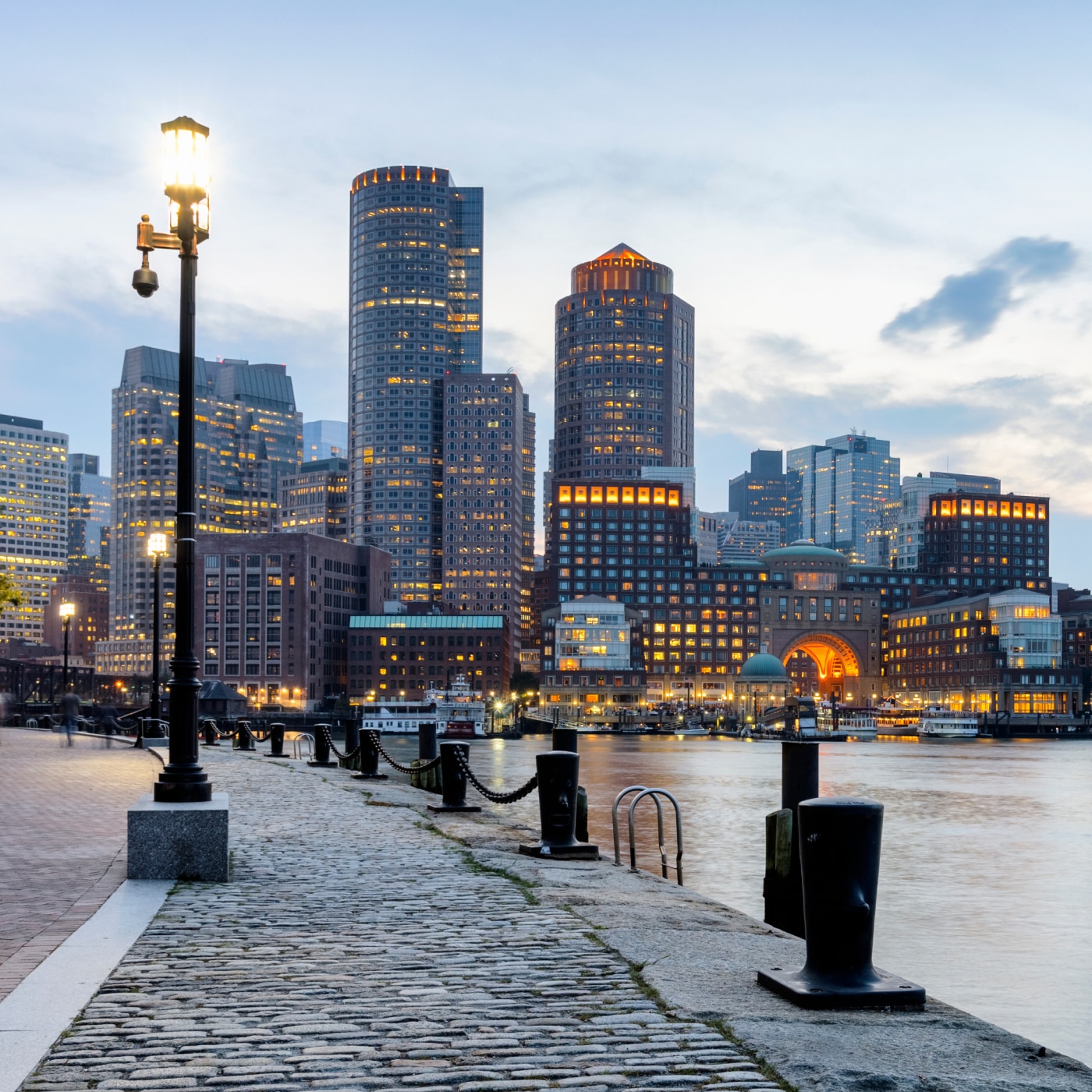 Waterfront view of downtown Boston at dusk with illuminated skyscrapers and cobblestone walkway.