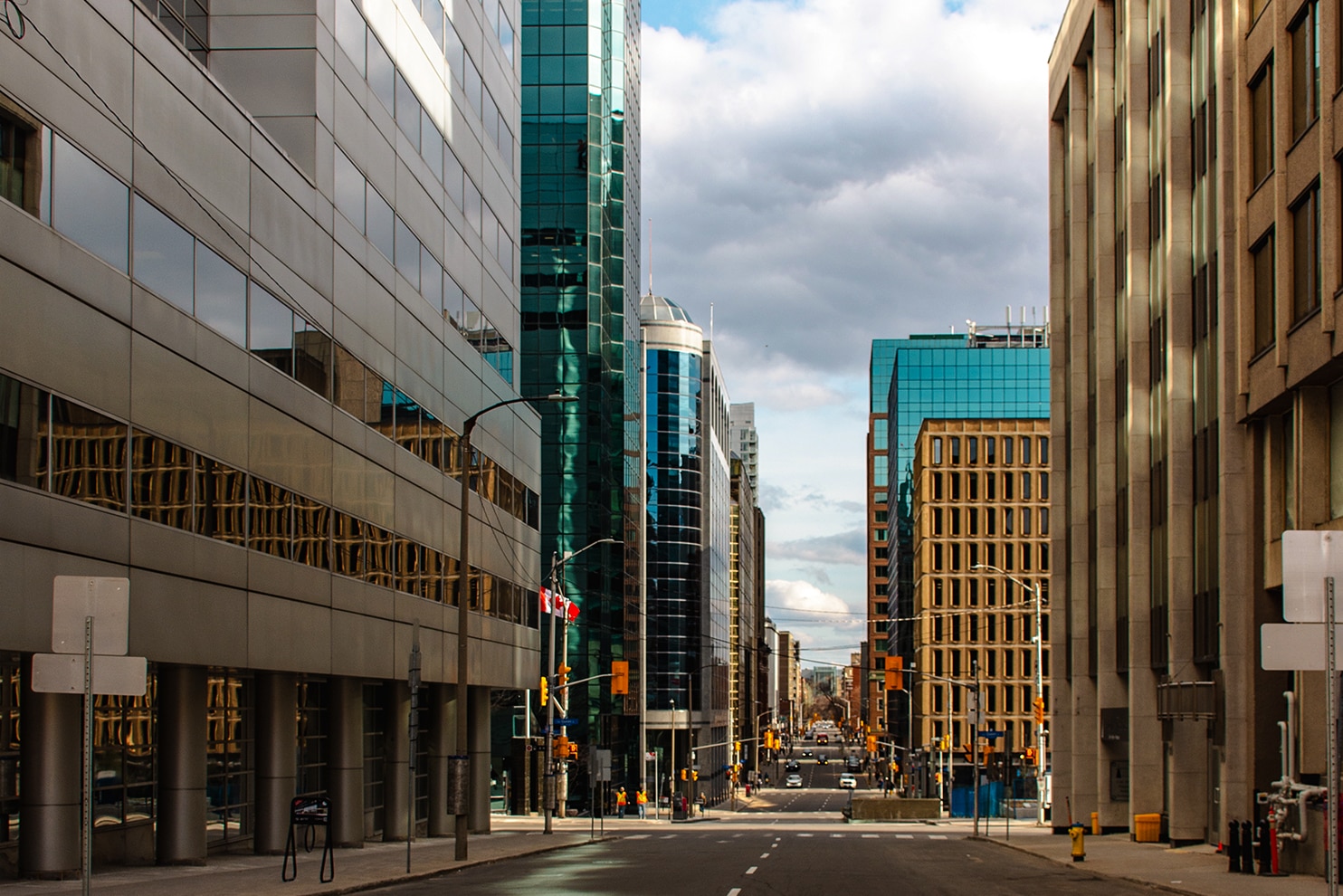 A quiet urban street lined with modern office buildings featuring glass and concrete facades. The road is slightly empty, with a few traffic lights and street signs visible. Reflections on the glass structures contrast with the rigid geometry of the surrounding architecture. The sky is partly cloudy, with patches of blue breaking through.