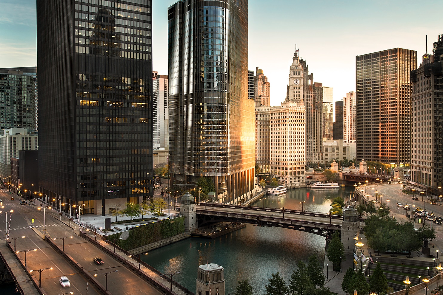 A stunning view of downtown Chicago at dusk, featuring the Chicago River and skyscrapers. The image captures illuminated bridges, modern glass buildings reflecting the fading sunlight and the bustling city streets with cars and pedestrians.