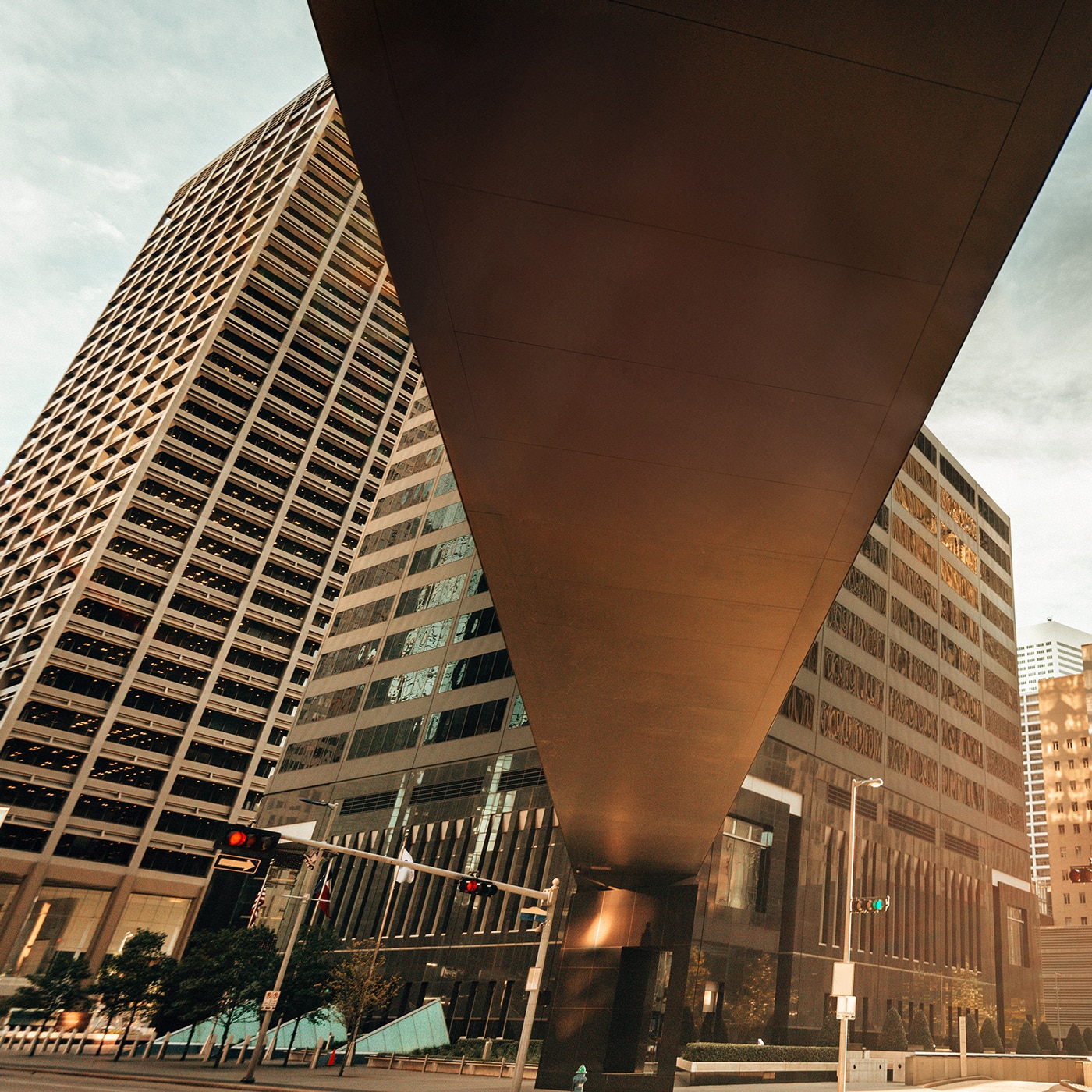 A dramatic urban scene captured from a low-angle perspective, featuring towering modern skyscrapers with reflective glass and geometric patterns. A massive overhead structure dominates the foreground. Traffic lights and street signs are visible at the base, adding to the dynamic city environment