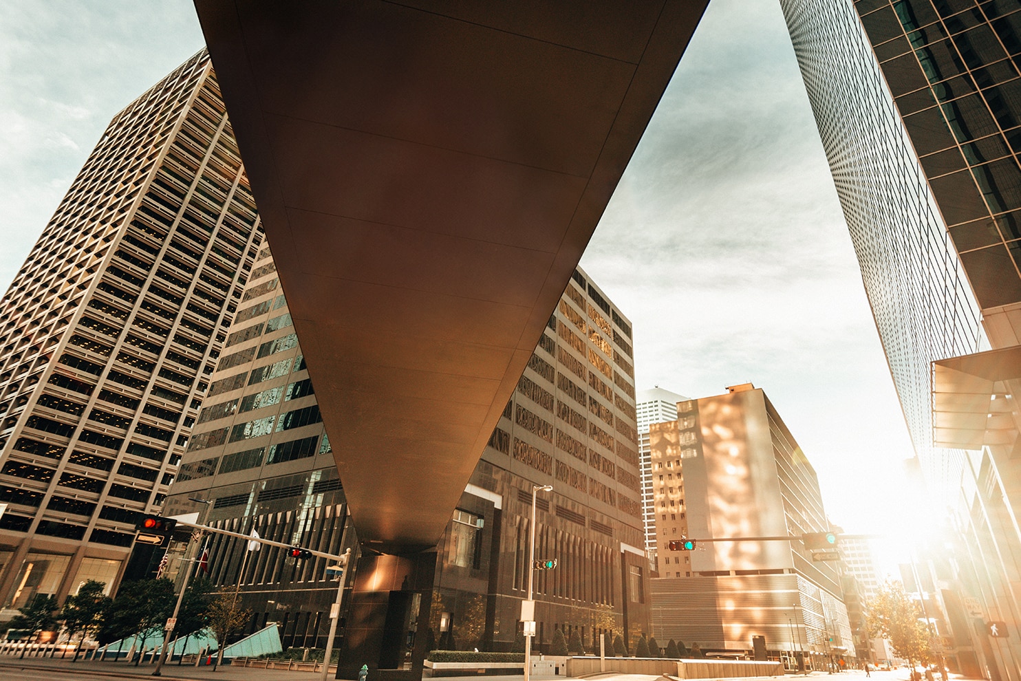 A dramatic urban scene captured from a low-angle perspective, featuring towering modern skyscrapers with reflective glass and geometric patterns. A massive overhead structure dominates the foreground. Traffic lights and street signs are visible at the base, adding to the dynamic city environment