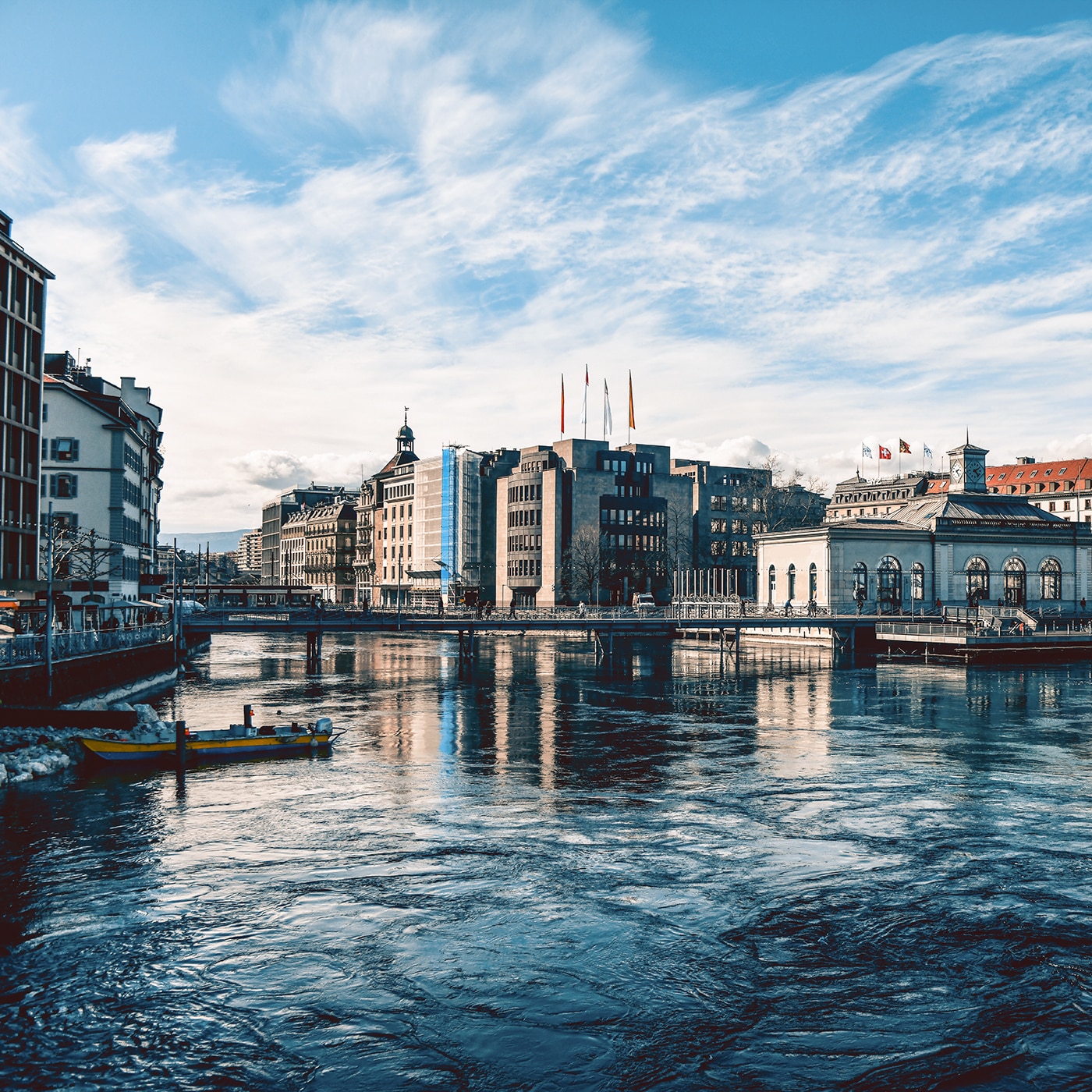 Waterfront buildings in Geneva with flags, a clock tower, and reflections on the Rhône River.