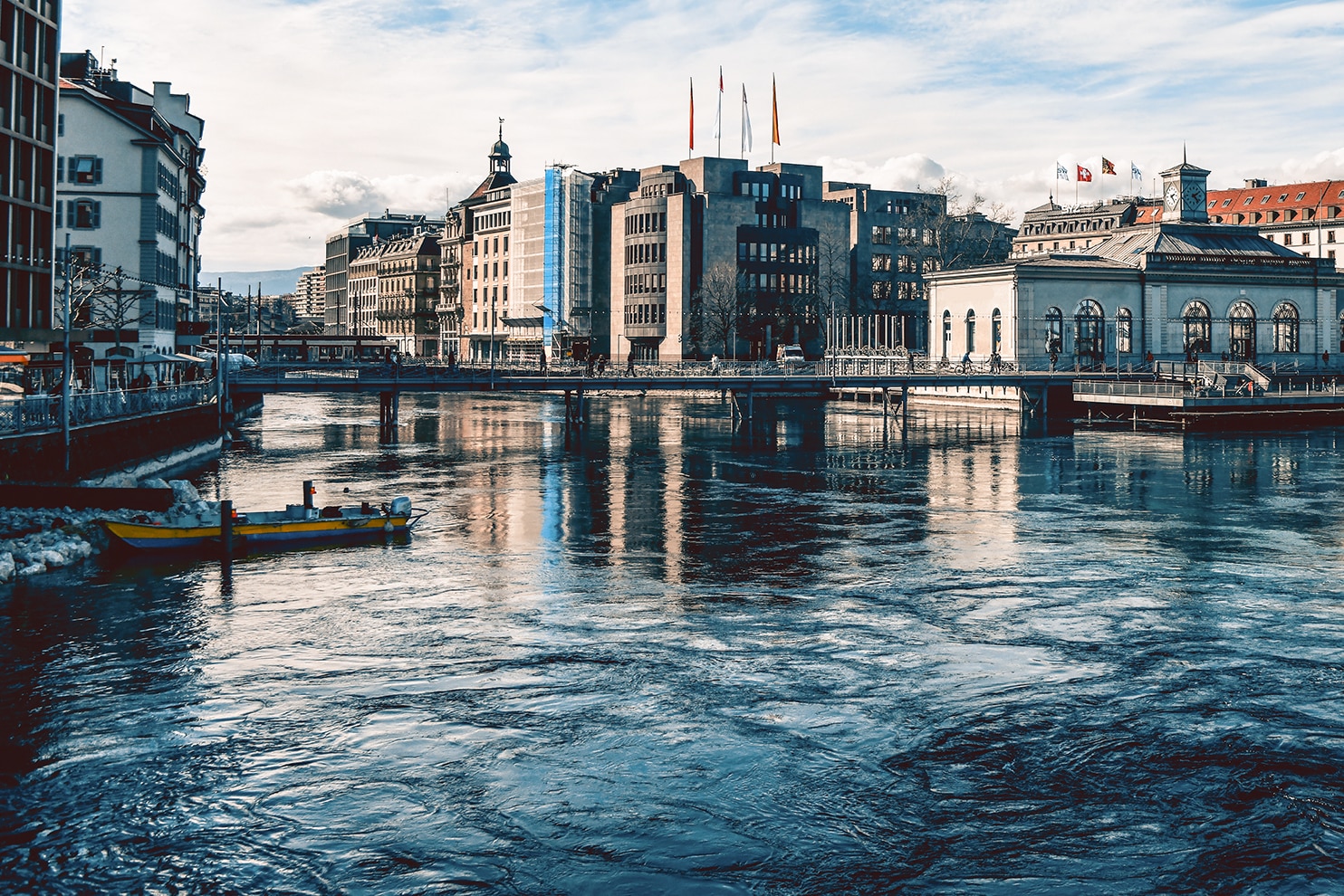 Waterfront buildings in Geneva with flags, a clock tower, and reflections on the Rhône River.