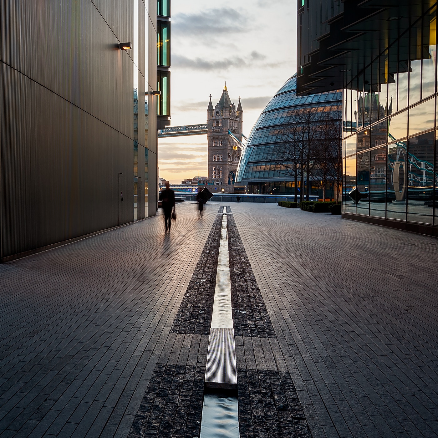 View through modern buildings towards Tower Bridge and City Hall at sunset in London.
