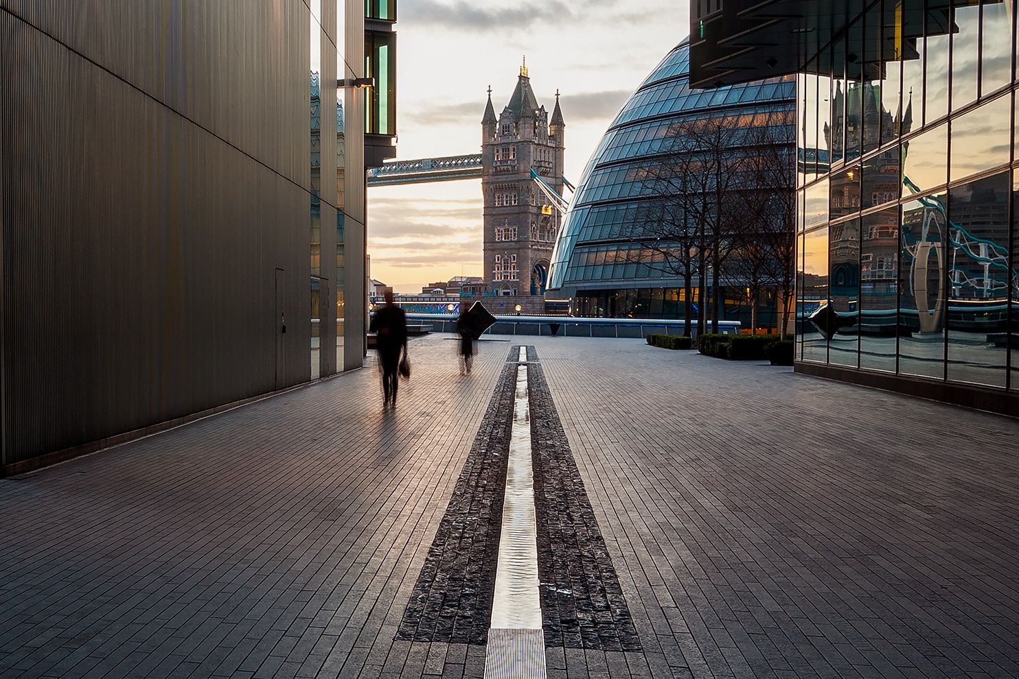 View through modern buildings towards Tower Bridge and City Hall at sunset in London.