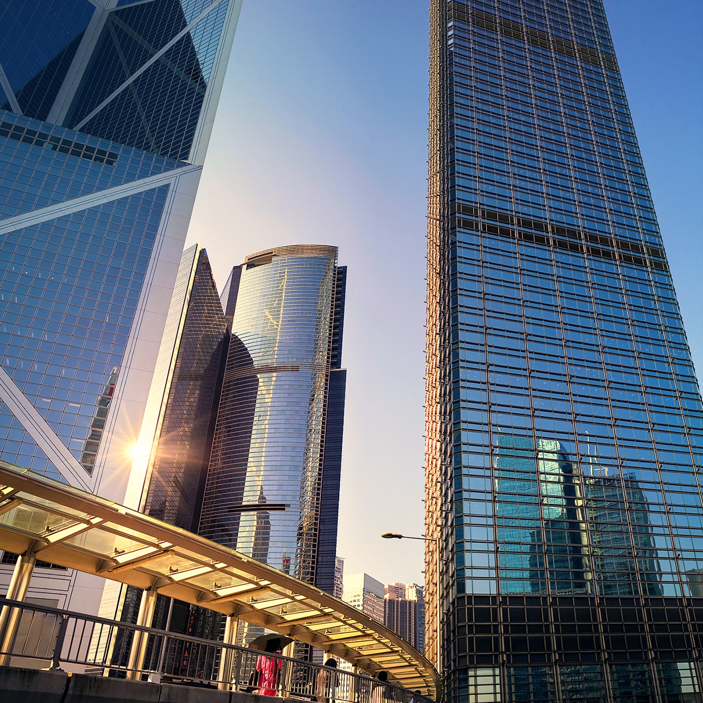 A dynamic low-angle view of modern skyscrapers in a financial district, reflecting sunlight on their glass facades. A curved pedestrian walkway in the foreground leads towards the towering buildings, with a few people walking along it. The deep blue sky contrasts with the sleek architecture, emphasizing the city's urban sophistication and fast-paced environment.