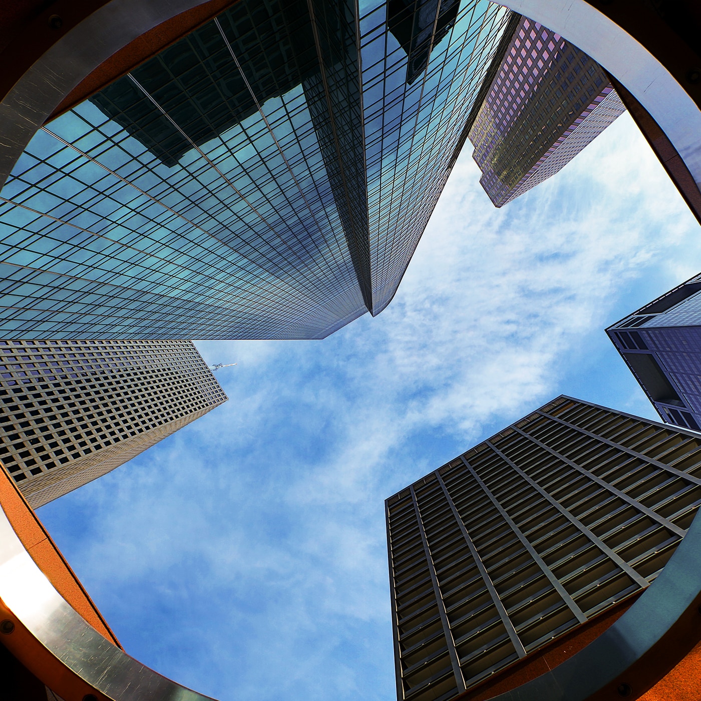 An urban scene with modern glass skyscrapers reflecting the sky and surrounding buildings. The foreground features a busy intersection with cars, pedestrians, and street signs. A large white building with a grid-like facade stands prominently, while a mix of contemporary and older architecture adds depth to the cityscape