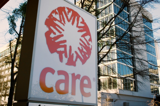 A large sign displaying the word "Care" with a stylized red emblem, set against the backdrop of a cityscape with glass buildings and trees.