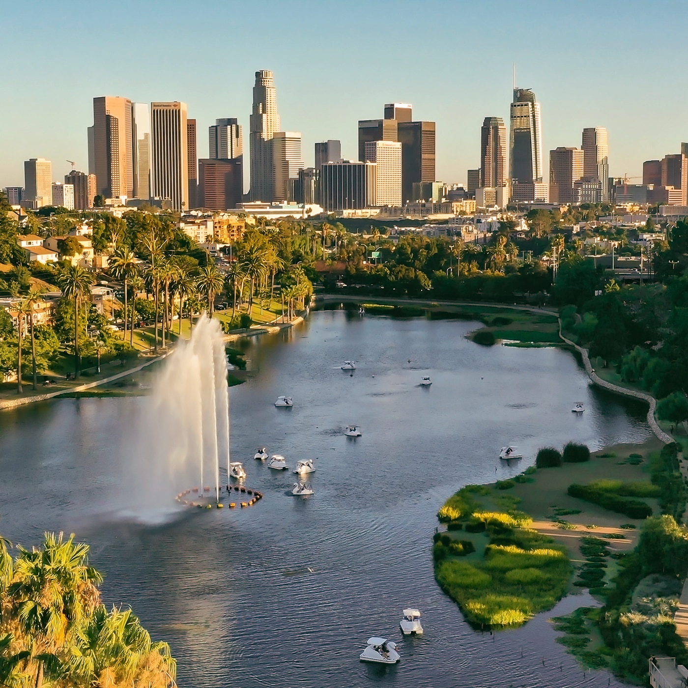 A scenic view of a large urban lake with a tall water fountain at its center, surrounded by lush greenery and palm trees. Small boats are scattered across the water. In the background, a towering city skyline with modern skyscrapers rises against a clear blue sky