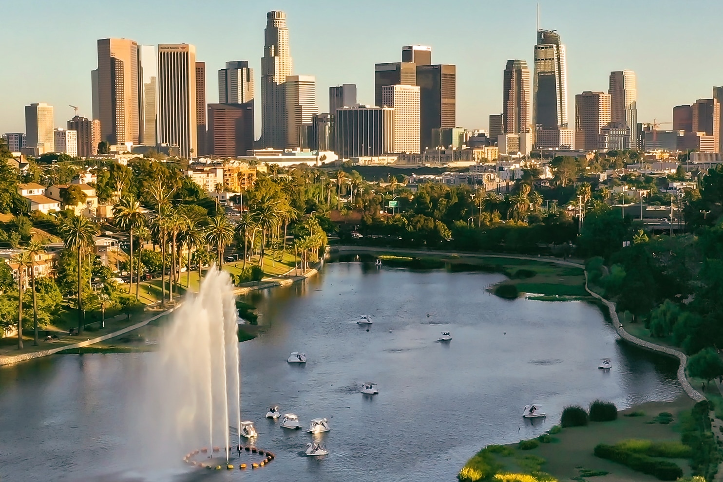 A scenic view of a large urban lake with a tall water fountain at its center, surrounded by lush greenery and palm trees. Small boats are scattered across the water. In the background, a towering city skyline with modern skyscrapers rises against a clear blue sky