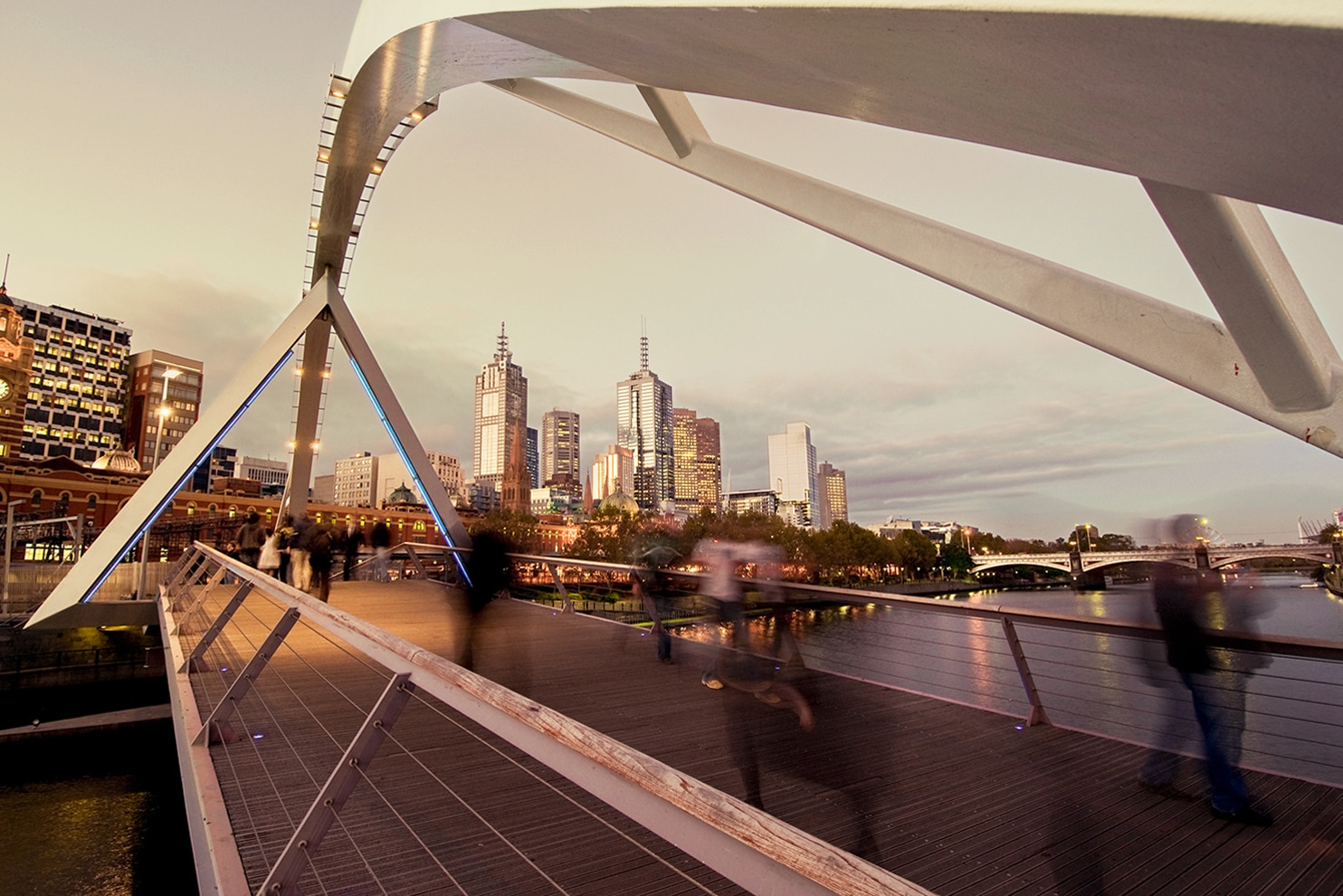 A cityscape of Melbourne, Australia, captured at dusk from a pedestrian bridge. The modern architectural design of the bridge frames the skyline, with illuminated high-rise buildings reflecting the fading daylight. Blurred figures of people walking across the bridge add a dynamic element, symbolizing the city's energy and movement. The Yarra River flows beneath, reflecting the warm lights of the city.
