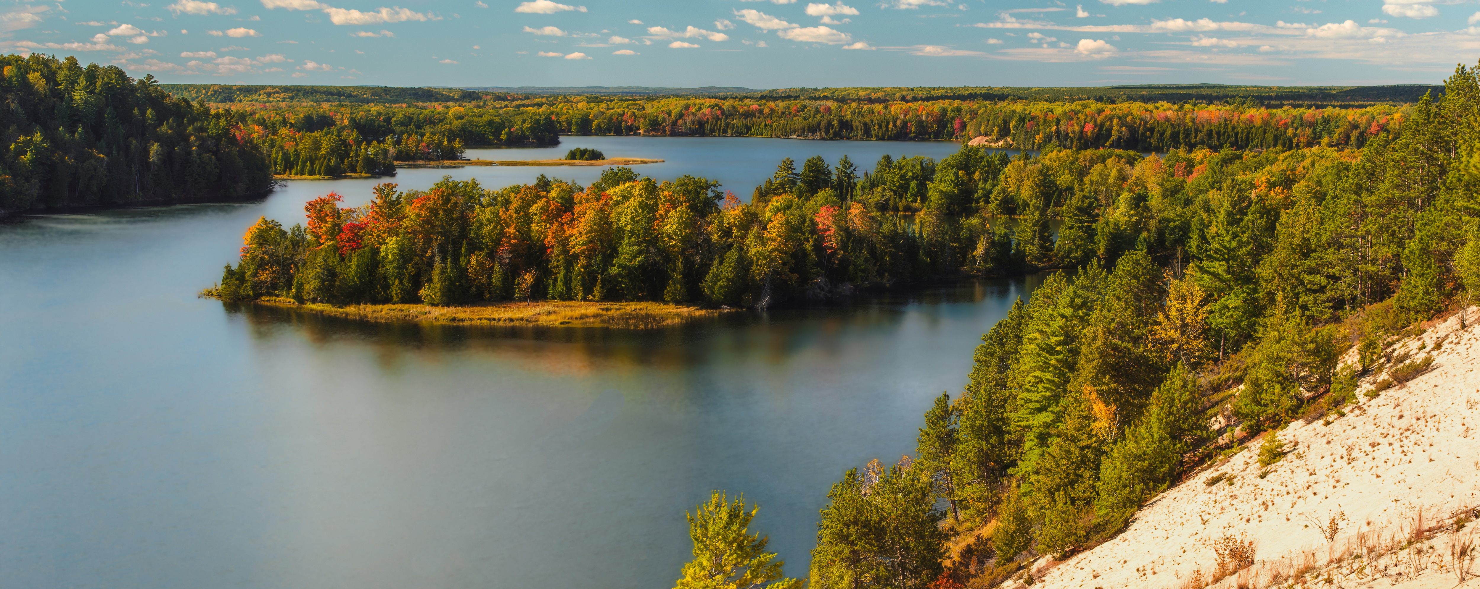 A scenic view of a calm lake surrounded by lush forests with vibrant autumn foliage. The image captures a sandy hillside in the foreground, overlooking the reflective water, with a backdrop of rolling wooded hills stretching to the horizon. The sky is blue with scattered white clouds