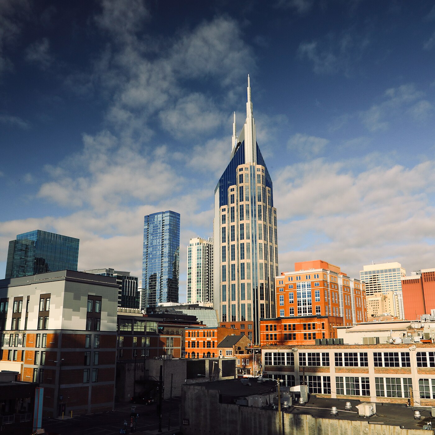 A cityscape featuring a modern skyline with a prominent, tall glass-clad skyscraper with twin spires. The surrounding buildings include a mix of contemporary high-rises and traditional brick structures. The sky is a deep blue with scattered white clouds.