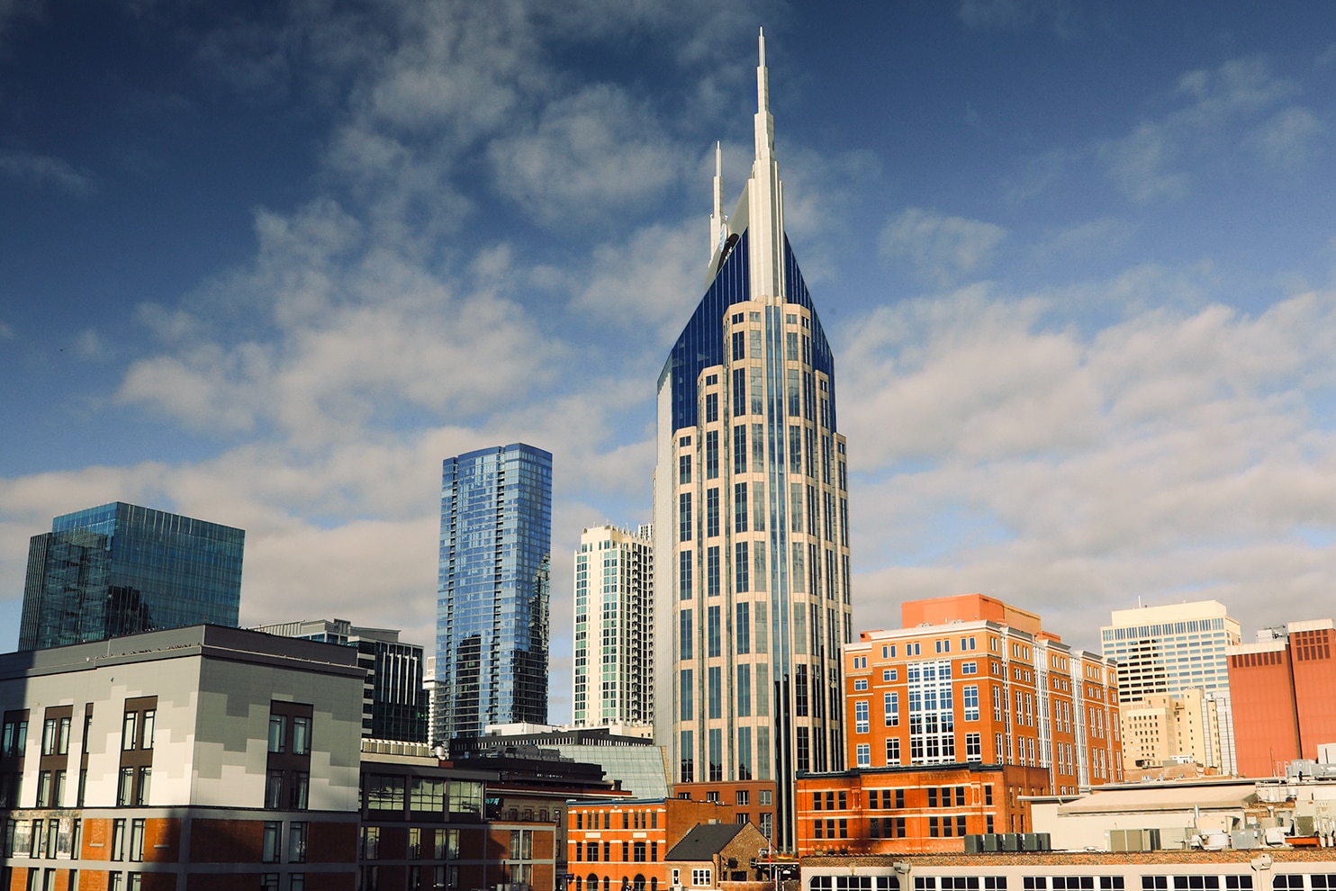 A cityscape featuring a modern skyline with a prominent, tall glass-clad skyscraper with twin spires. The surrounding buildings include a mix of contemporary high-rises and traditional brick structures. The sky is a deep blue with scattered white clouds.