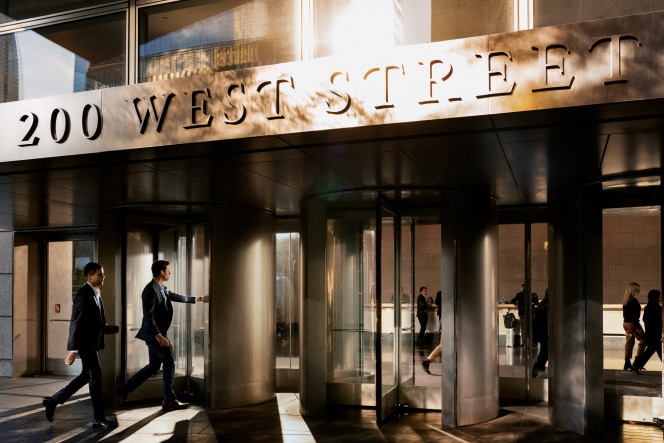 Two professionally dressed men in business suits enter a modern office building at 200 West Street through revolving glass doors. The warm glow of sunlight casts long shadows on the ground, highlighting the sleek architecture of the entrance. Reflections of people inside the building are visible through the glass