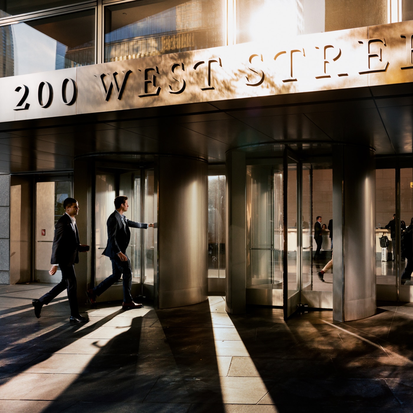 Two professionally dressed men in business suits enter a modern office building at 200 West Street through revolving glass doors. The warm glow of sunlight casts long shadows on the ground, highlighting the sleek architecture of the entrance. Reflections of people inside the building are visible through the glass