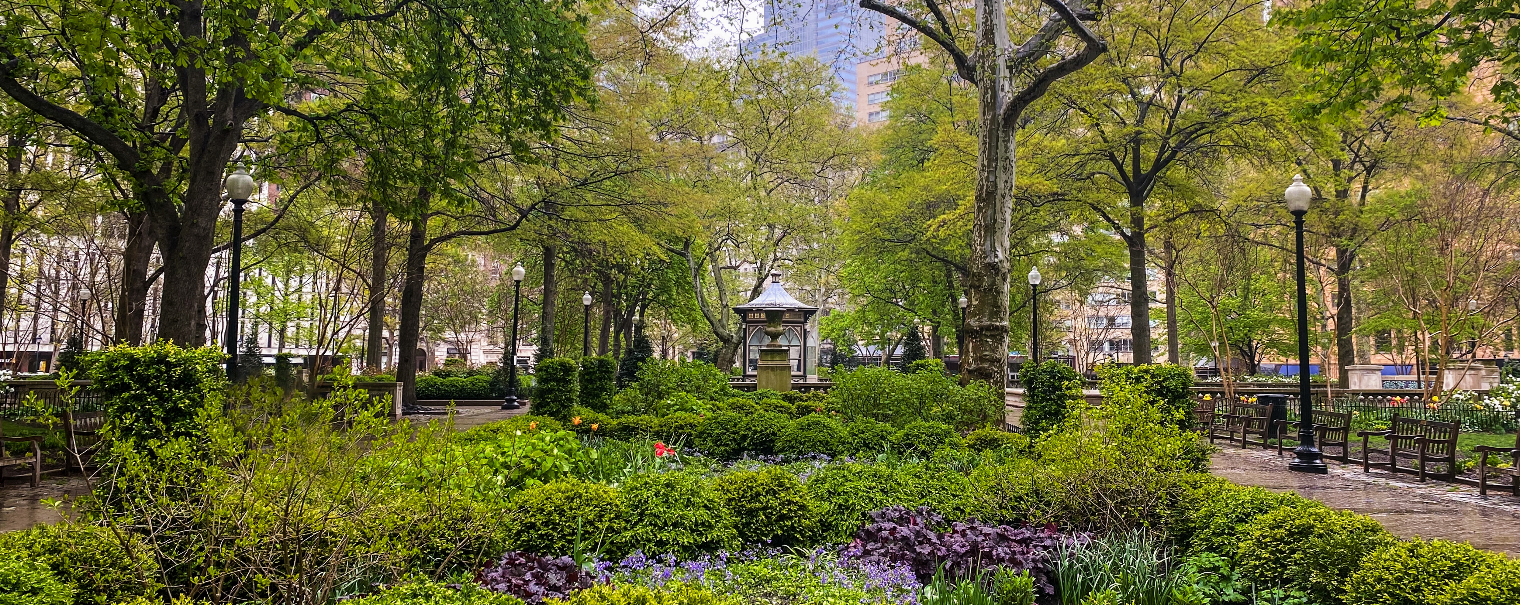 Lush green city park with benches, lampposts, flower beds, and a small pavilion surrounded by trees and buildings.