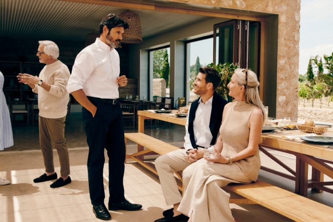 A man in a white shirt and dark trousers chats with a smiling couple seated at an outdoor dining area, surrounded by a stylish open space with a set wooden table.