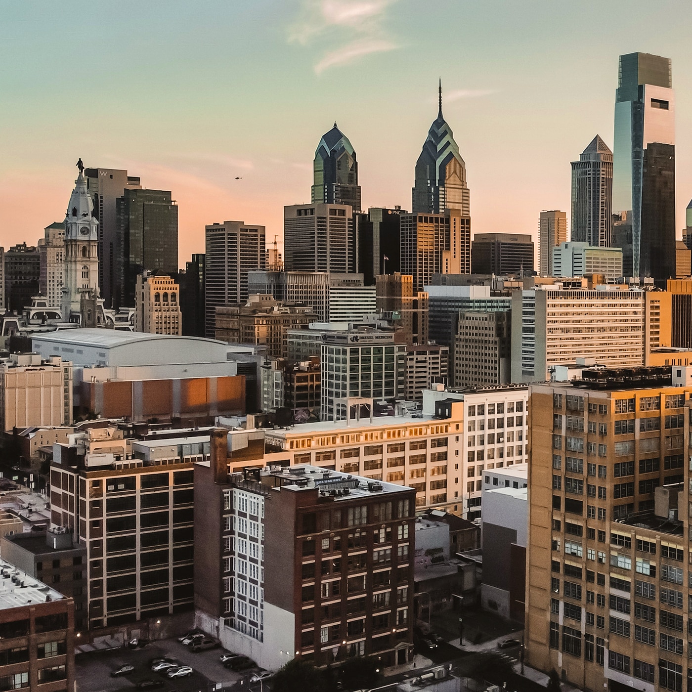 A cityscape of Philadelphia at sunset, featuring a mix of modern skyscrapers and historic buildings. The skyline includes Comcast Center and Liberty Place towers, with the white clock tower of City Hall standing prominently in the foreground. The warm glow of the setting sun casts a golden hue on the buildings, highlighting their architectural details.