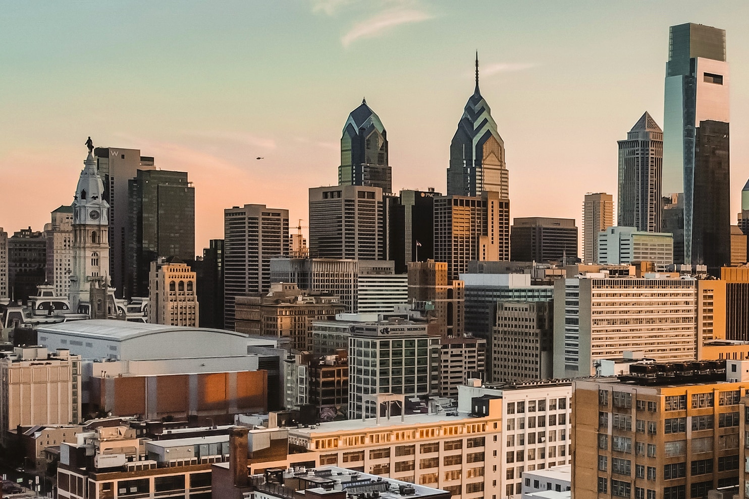A cityscape of Philadelphia at sunset, featuring a mix of modern skyscrapers and historic buildings. The skyline includes Comcast Center and Liberty Place towers, with the white clock tower of City Hall standing prominently in the foreground. The warm glow of the setting sun casts a golden hue on the buildings, highlighting their architectural details.