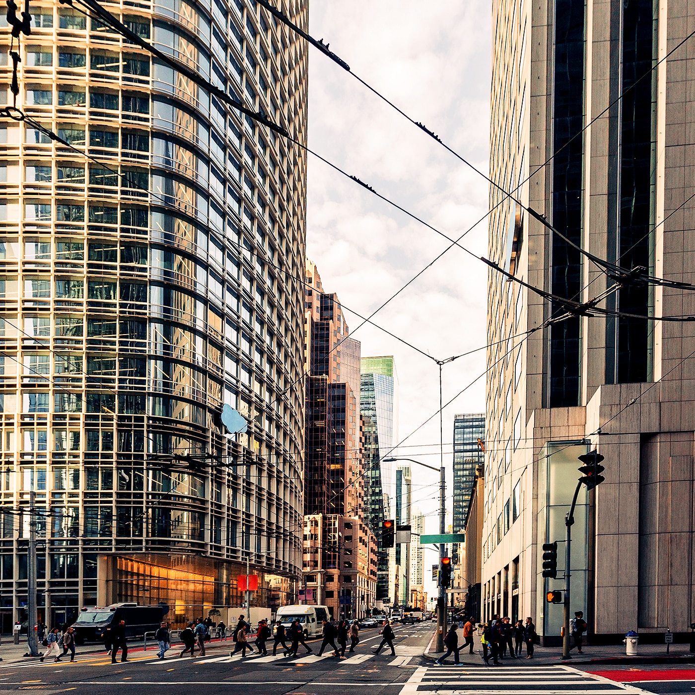 A busy city intersection surrounded by towering modern skyscrapers with reflective glass facades. Pedestrians cross the street while vehicles navigate the urban environment. Overhead, a network of cables crisscrosses the sky. The bright sky contrasts with the dense architecture