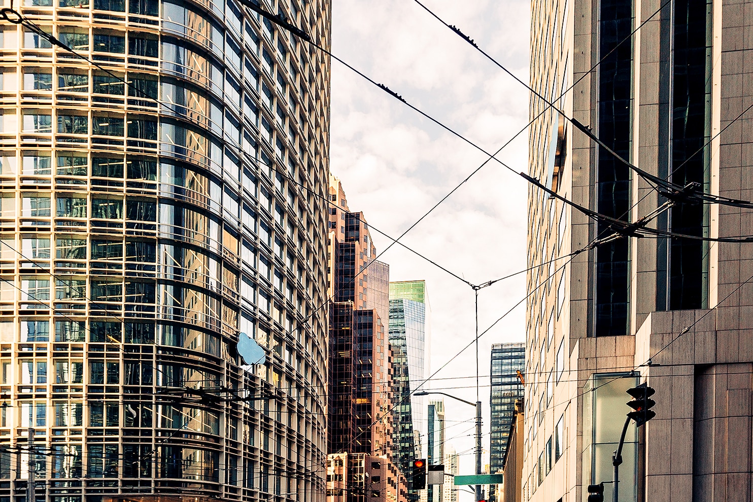 A busy city intersection surrounded by towering modern skyscrapers with reflective glass facades. Pedestrians cross the street while vehicles navigate the urban environment. Overhead, a network of cables crisscrosses the sky. The bright sky contrasts with the dense architecture