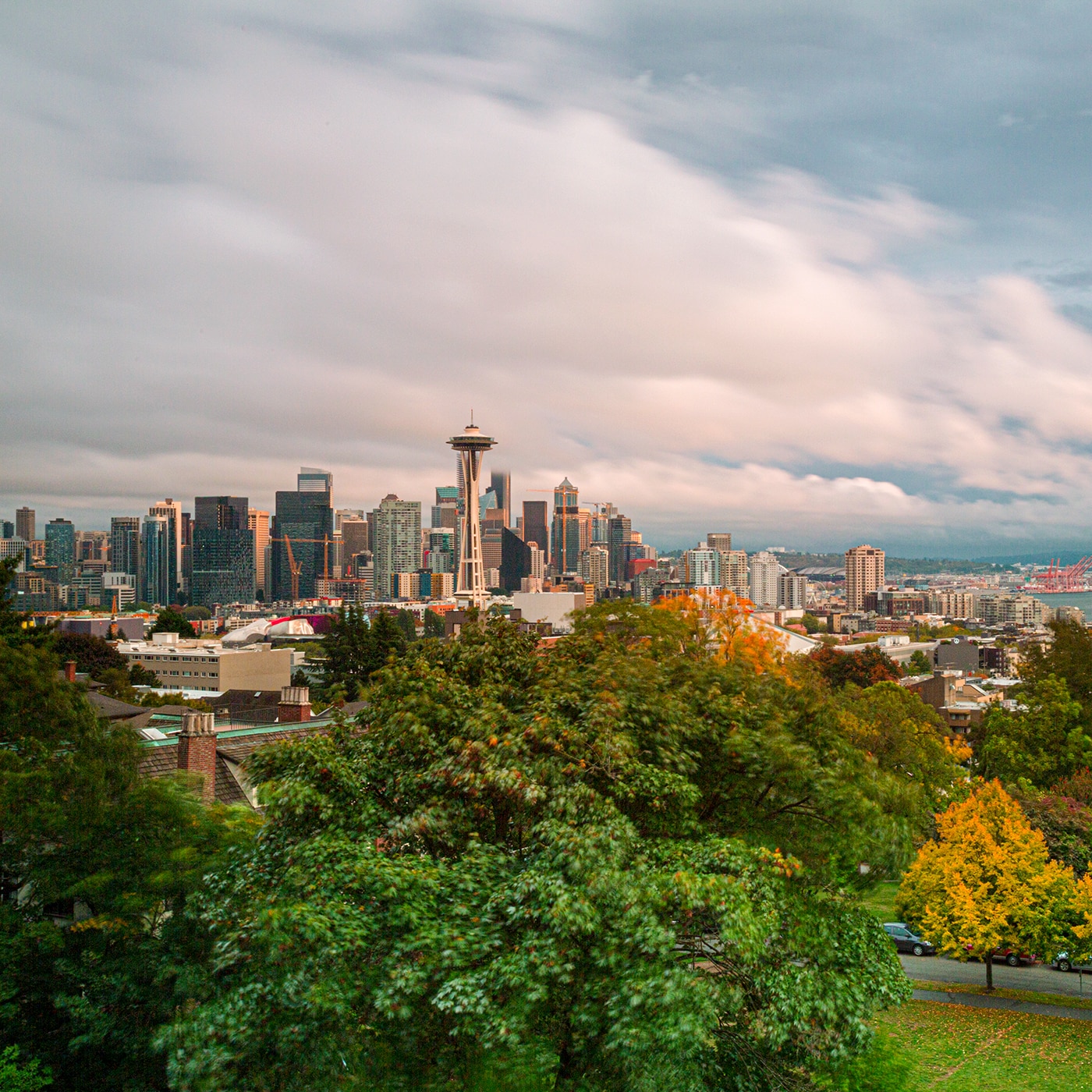 A cityscape of Seattle featuring the Space Needle standing among modern skyscrapers. The view is framed by lush green trees in the foreground, with a dramatic cloudy sky above. The city's urban landscape extends into the distance, with glimpses of waterfront areas and distant hills