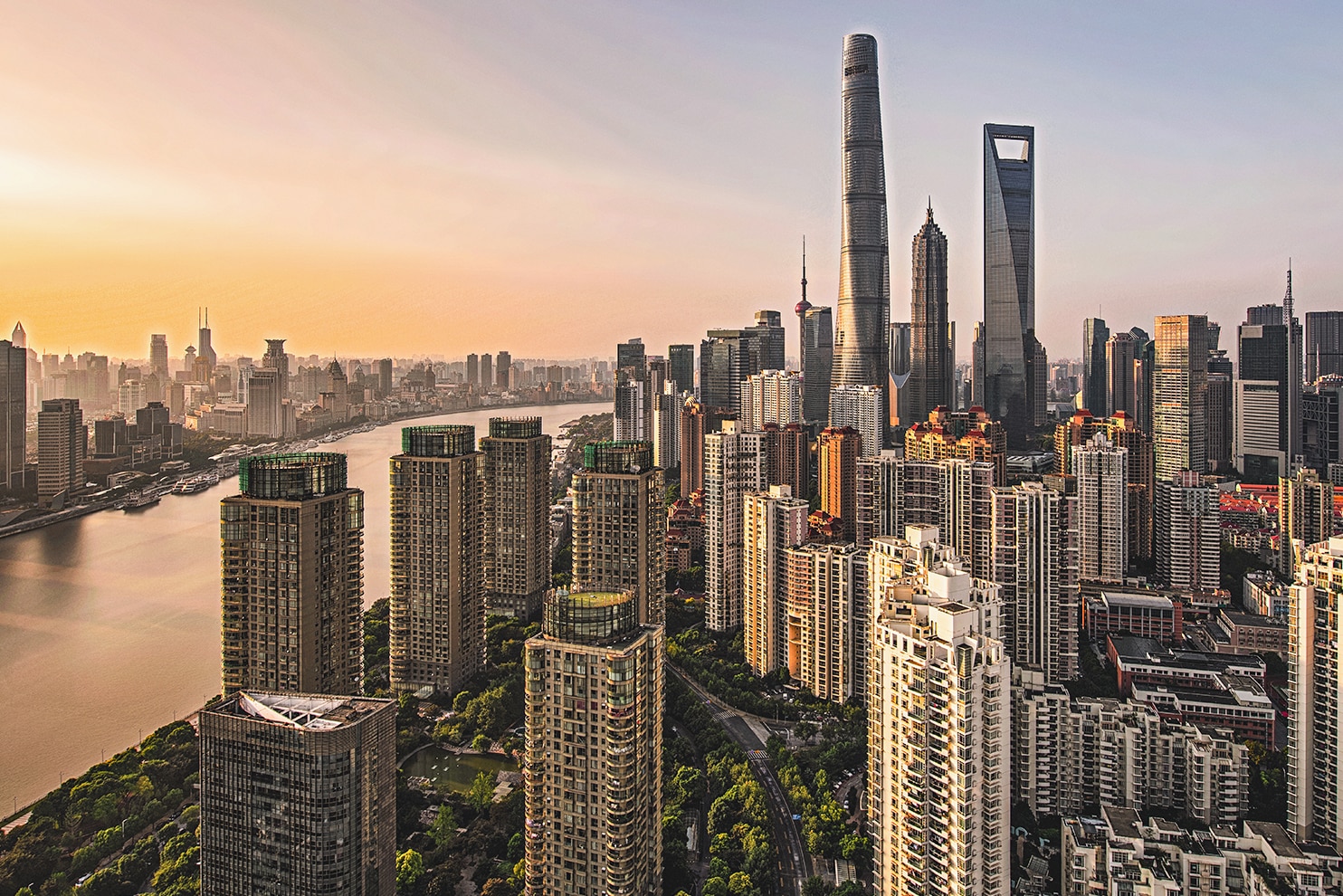A cityscape of a modern metropolis with numerous high-rise buildings, including skyscrapers. A river runs through the city, and the skyline is illuminated by soft sunlight. The architecture is a mix of residential and commercial structures, with green spaces visible among the buildings.