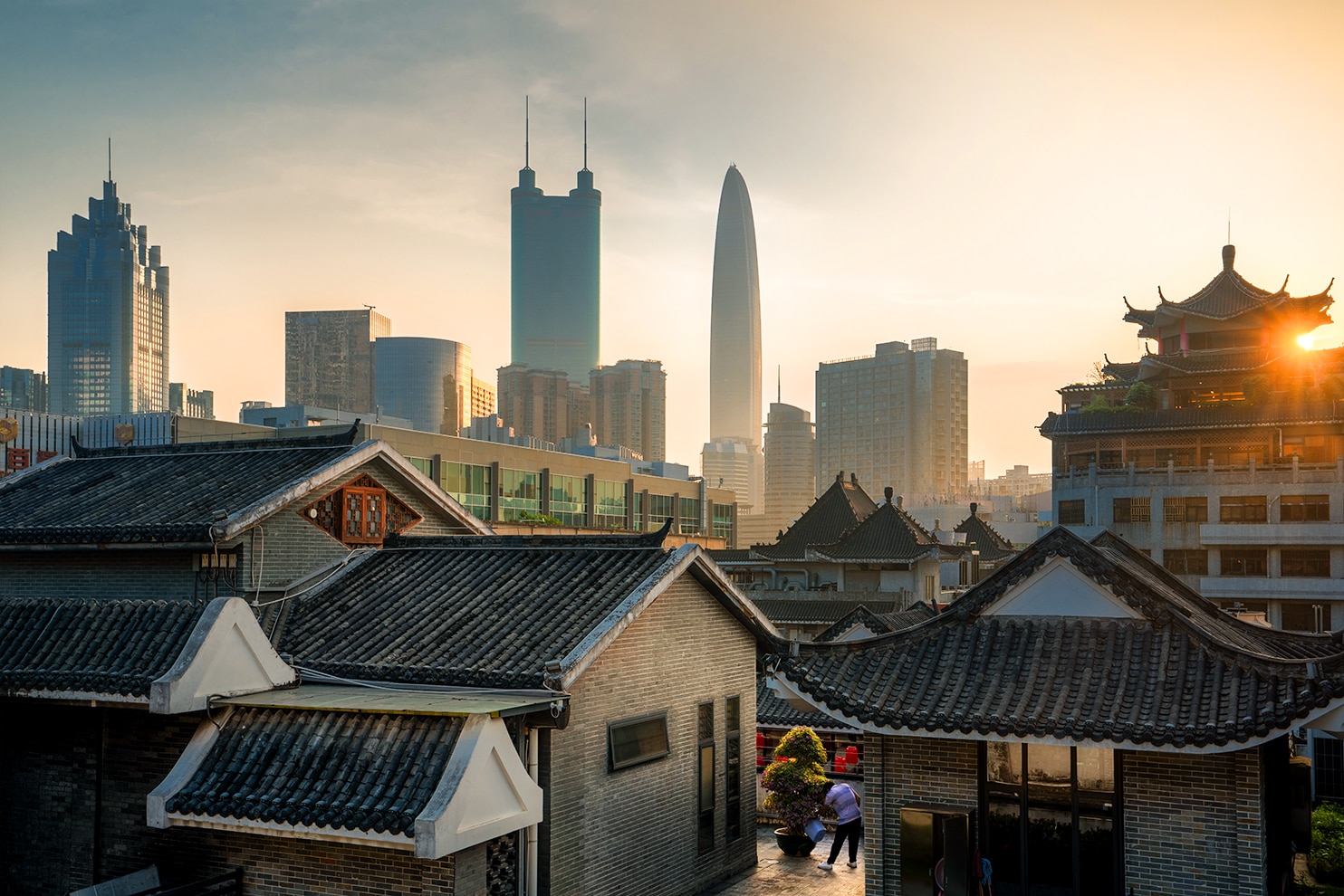 A cityscape at sunset showcasing the contrast between traditional Chinese architecture with dark tiled rooftops in the foreground and modern skyscrapers in the background. The setting sun casts a warm glow on the historic buildings, highlighting intricate roof details, while the glass skyscrapers, including a distinctive tall, sleek tower, rise in the skyline. A person is seen tending to plants in a courtyard, adding a human touch to the dynamic urban scene.