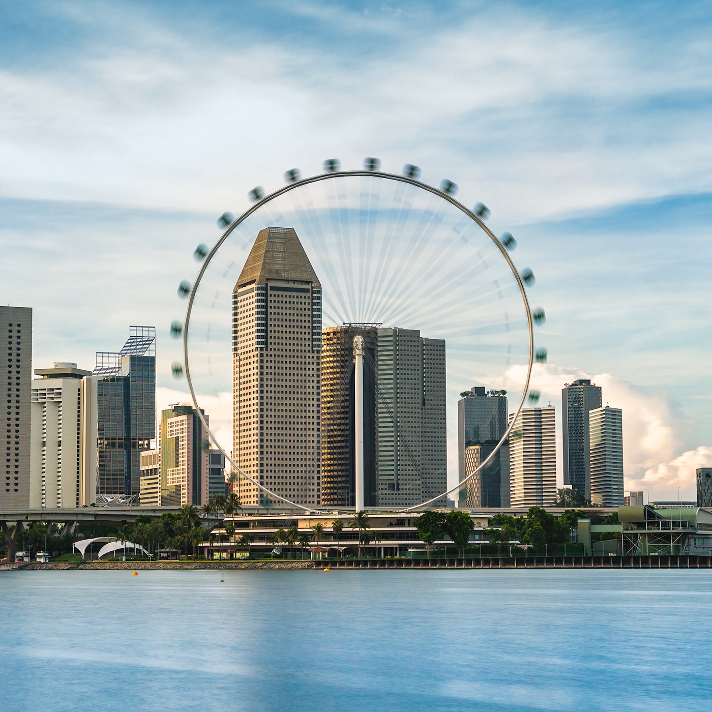 A view of the Singapore skyline featuring the Singapore Flyer, a large observation wheel, set against modern skyscrapers. The cityscape is reflected in the calm waters in the foreground, while the sky is a blend of soft blues and wispy clouds. The composition highlights the harmony between urban architecture and waterfront scenery, capturing the vibrancy and elegance of Singapore.