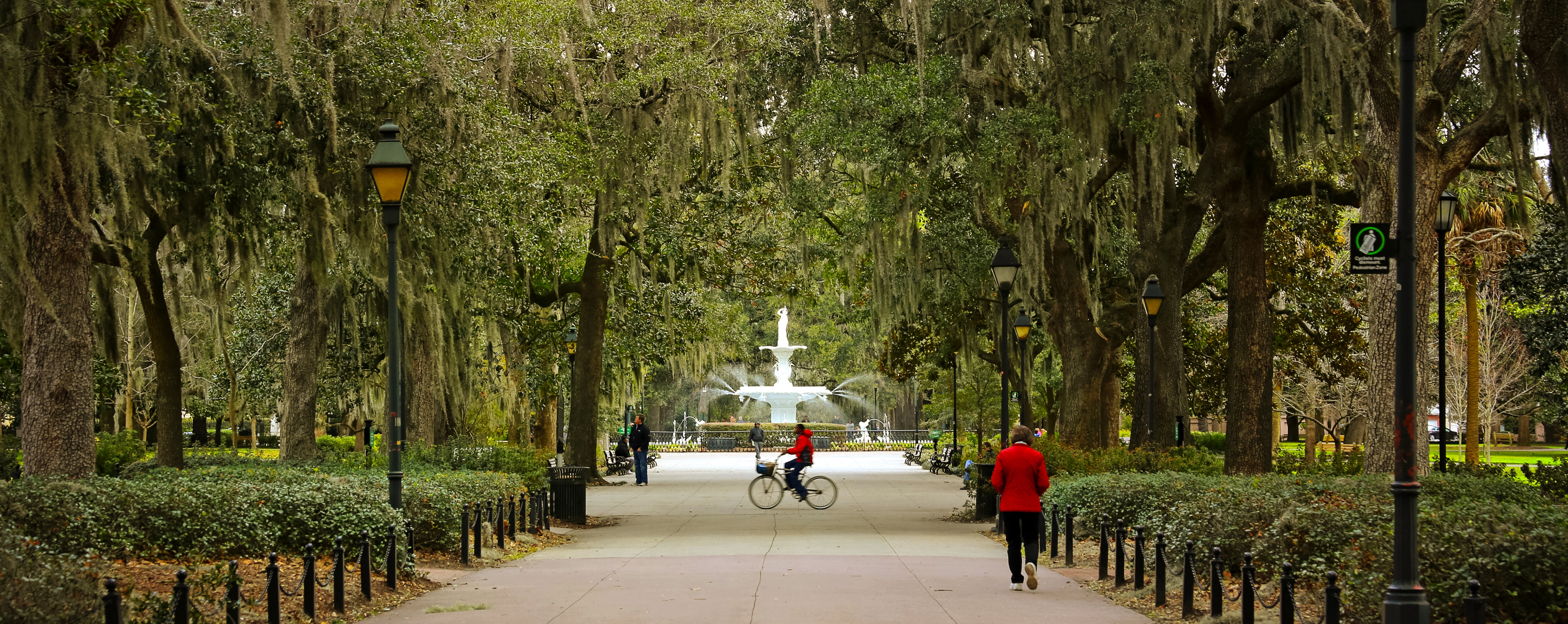 A scenic park pathway lined with tall oak trees draped in Spanish moss leads to a grand white fountain in the distance. Several people are walking, cycling, and enjoying the surroundings. Traditional lampposts line the path and lush greenery
