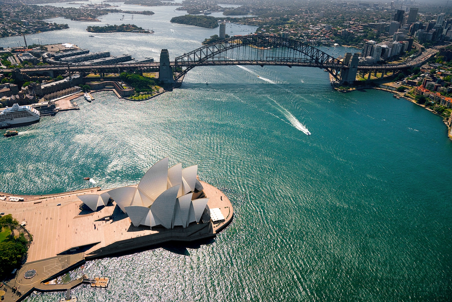 An aerial view of Sydney Harbour, featuring the Sydney Opera House with its distinctive white sail-like structures and the Sydney Harbour Bridge. The deep blue waters of the harbor are dotted with boats leaving white trails as they move. The city skyline, lush green parks, and surrounding urban areas stretch into the background, highlighting Sydney's vibrant waterfront.