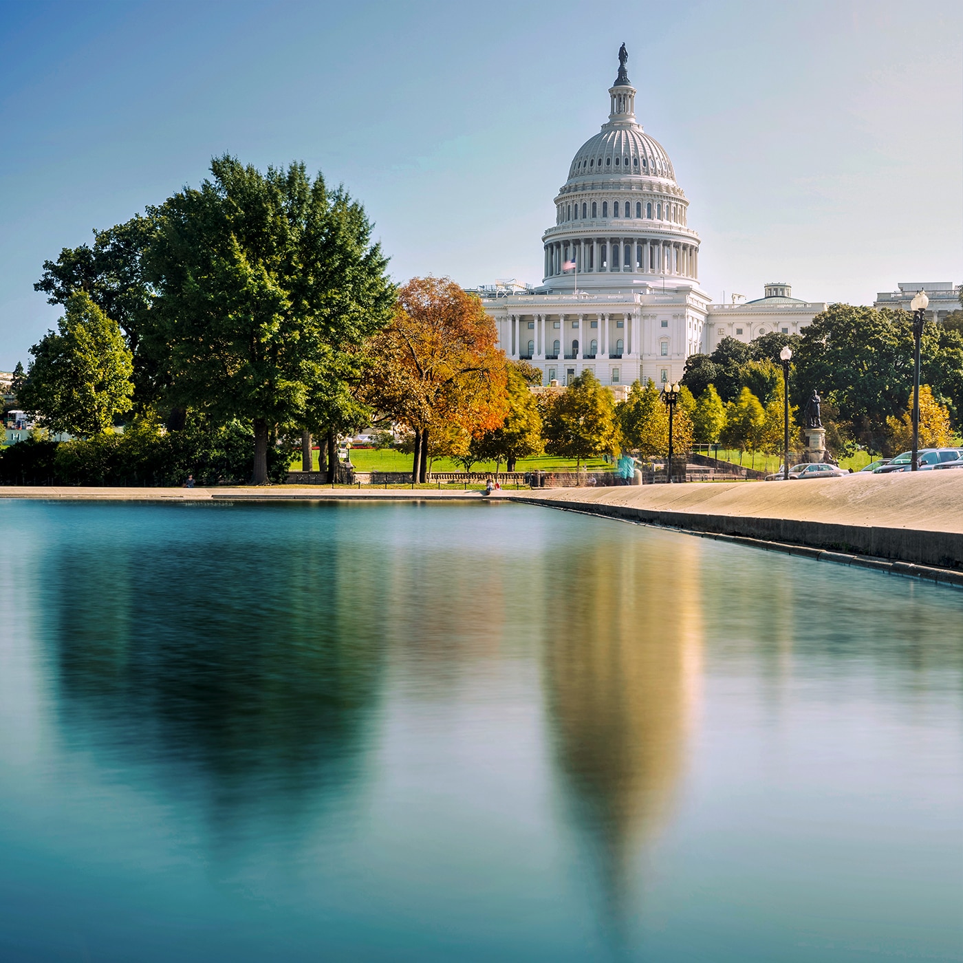 The United States Capitol building in Washington, D.C., with its dome, stands against a clear blue sky. In the foreground, the Capitol Reflecting Pool mirrors the surrounding trees and architecture. The lush greenery and autumn-colored foliage add vibrancy to the setting