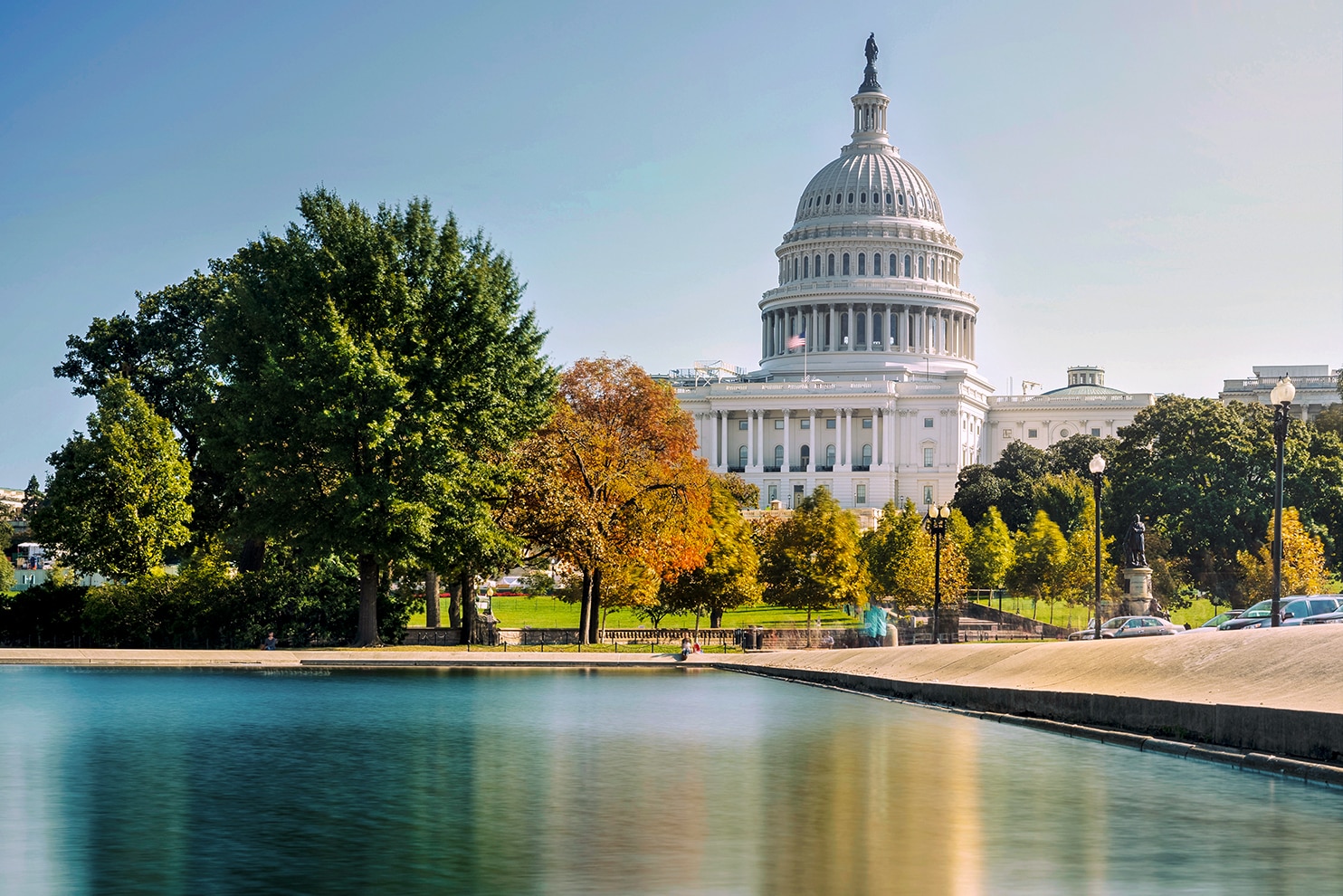 The United States Capitol building in Washington, D.C., with its dome, stands against a clear blue sky. In the foreground, the Capitol Reflecting Pool mirrors the surrounding trees and architecture. The lush greenery and autumn-colored foliage add vibrancy to the setting