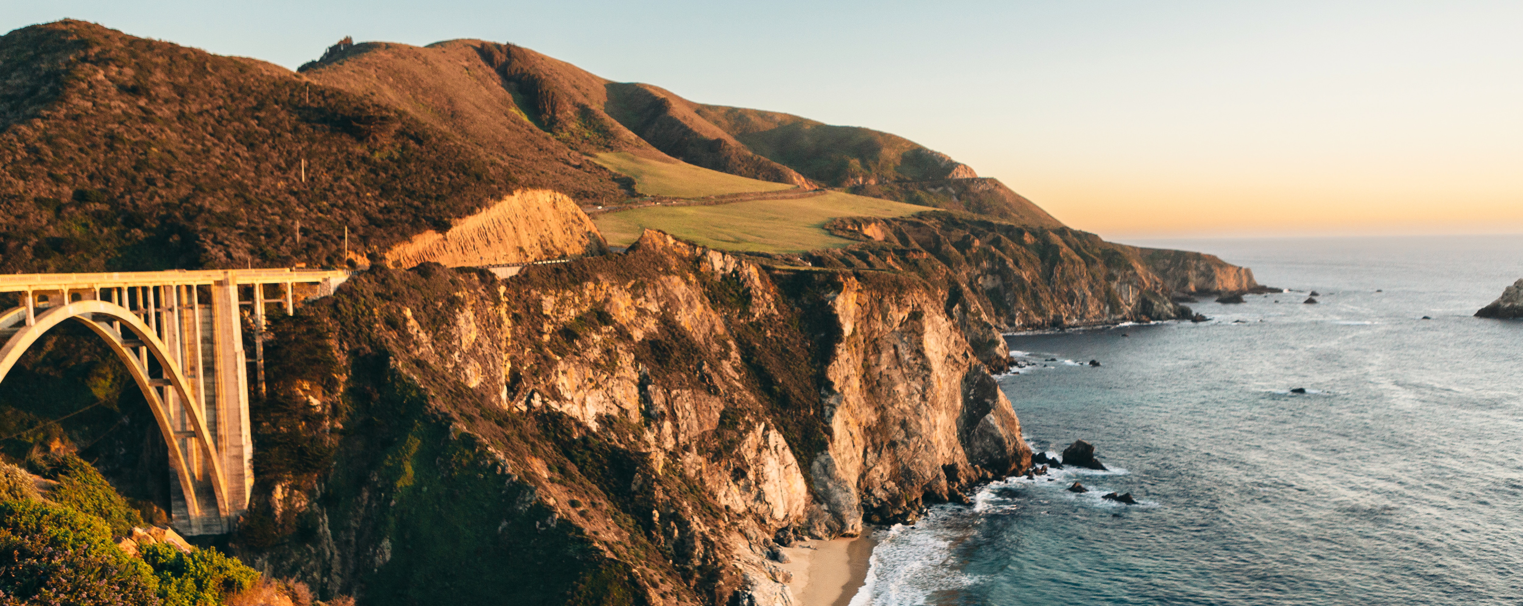 A coastal landscape at sunset, featuring a rugged cliffside meeting the deep blue ocean. An arched bridge spans a canyon, connecting a winding road along the coastline. Rolling green hills in the background contrast with the rocky cliffs, while waves lap against the shoreline below. The sky transitions from warm golden hues to cooler blues