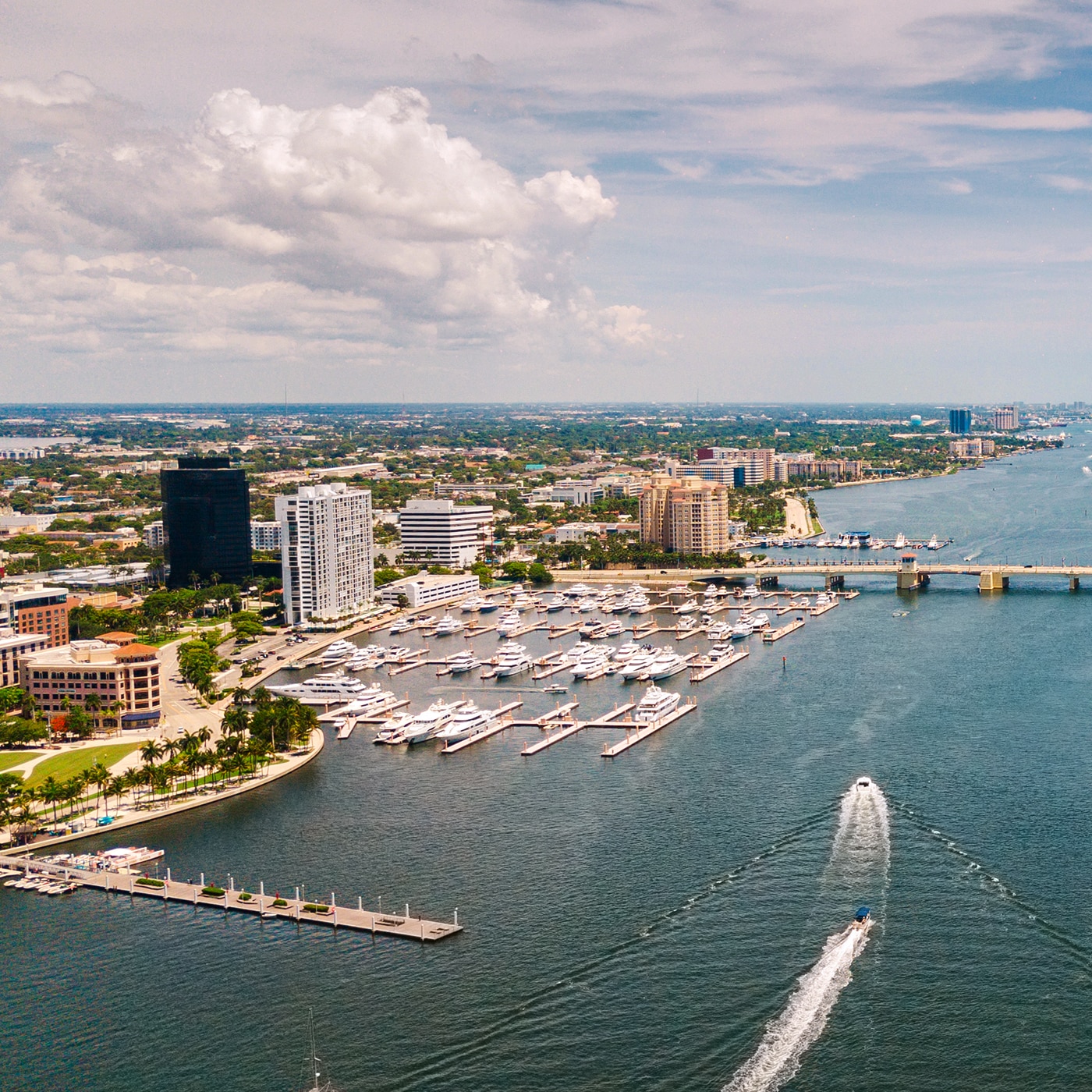 Aerial view of a coastal city with a marina filled with yachts and boats, a bridge spanning the waterway, and a speedboat creating a wake as it moves through the water. The city skyline features a mix of modern high-rises and low-rise buildings under a partly cloudy sky