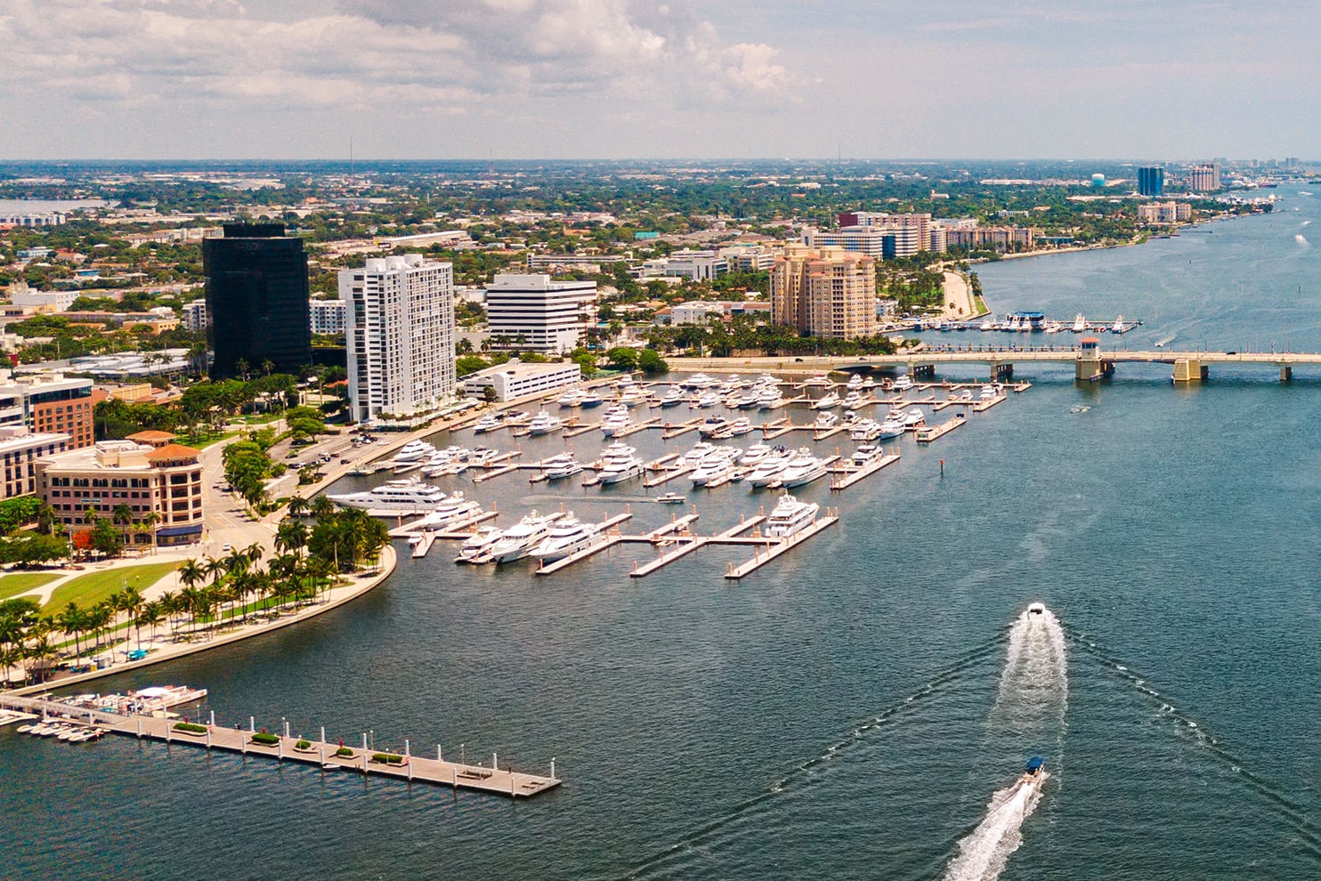 Aerial view of a coastal city with a marina filled with yachts and boats, a bridge spanning the waterway, and a speedboat creating a wake as it moves through the water. The city skyline features a mix of modern high-rises and low-rise buildings under a partly cloudy sky