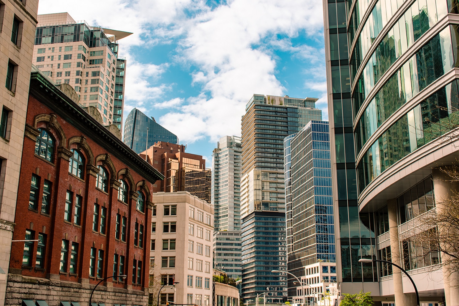 A cityscape view featuring a mix of historic and modern architecture. A red brick building with arched windows contrasts with sleek glass and steel skyscrapers in the background. A curved glass structure reflects the urban surroundings. The sky is partly cloudy with patches of blue.