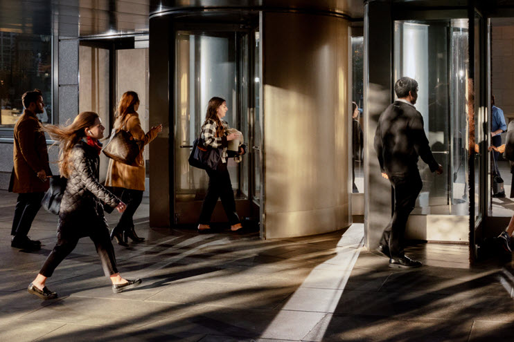 People entering and exiting a building through revolving doors in the early morning light.