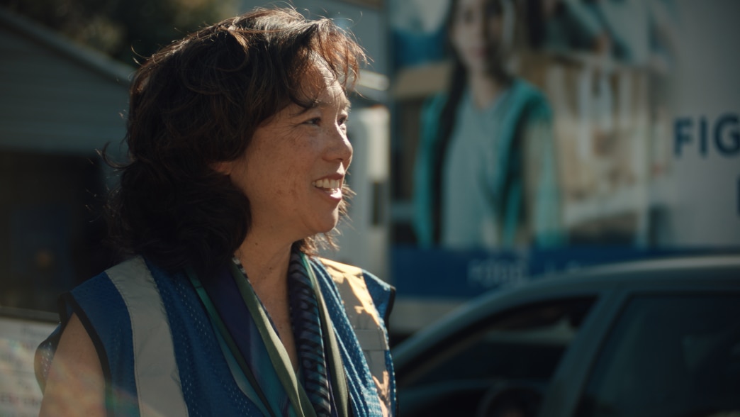 Kathy Kwan wears a reflective safety vest and a scarf smiles while standing outdoors. 