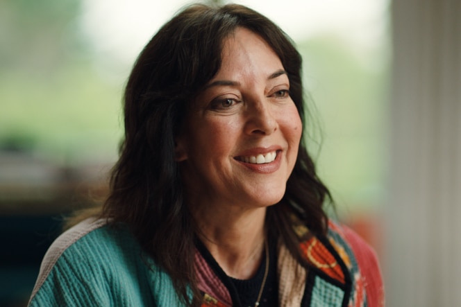 Leslie Blodgett sits on a vibrant yellow couch, wearing a colorful jacket, with a bright window, greenery, and decorative pillows in the background.