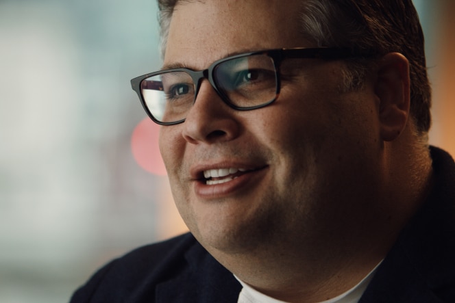 RJ Melman wears a dark blazer and smiles while speaking in a softly lit indoor setting.