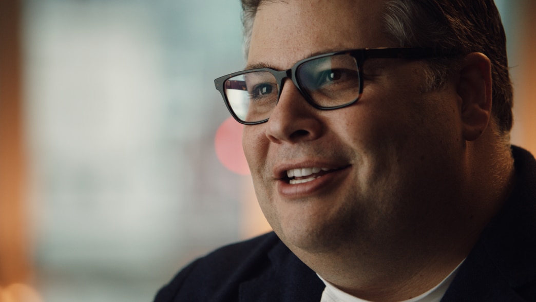 RJ Melman wears a dark blazer and smiles while speaking in a softly lit indoor setting.